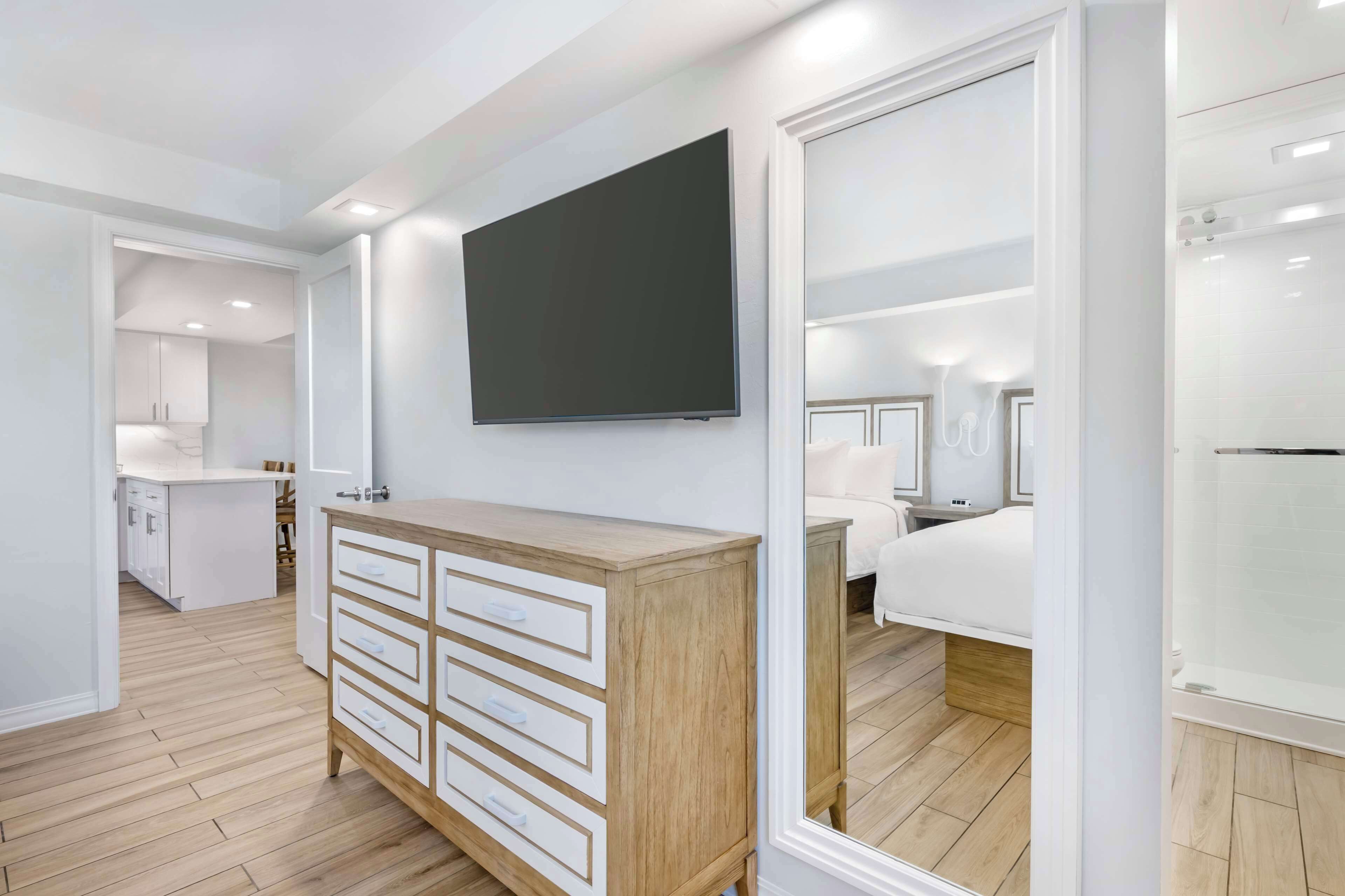 A modern hotel room with a wooden dresser, a flat-screen TV, a large mirror, and a separate bedroom area.