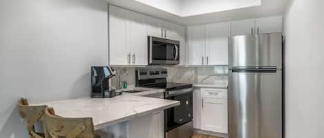 A modern kitchen with white cabinets, stainless steel appliances, a marble countertop, and wooden chairs.