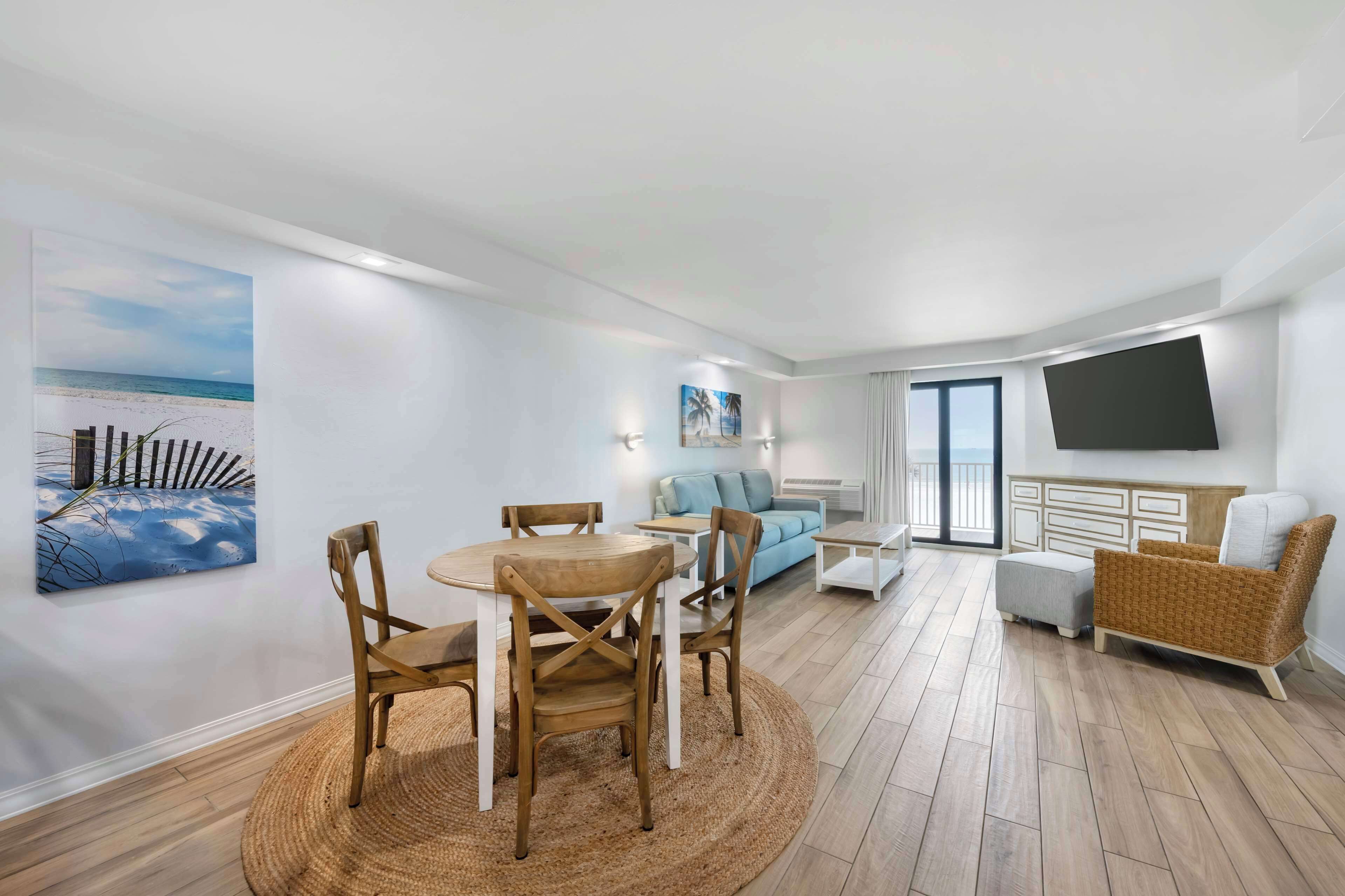 A modern living room with a round wooden table and chairs, a light blue sofa, a wicker armchair, and a flat-screen TV mounted on the wall.
