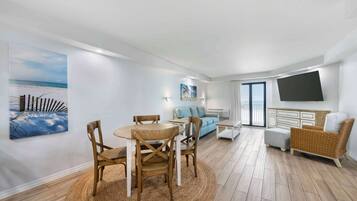 A modern living room with a round wooden table and chairs, a light blue sofa, a wicker armchair, and a flat-screen TV mounted on the wall.