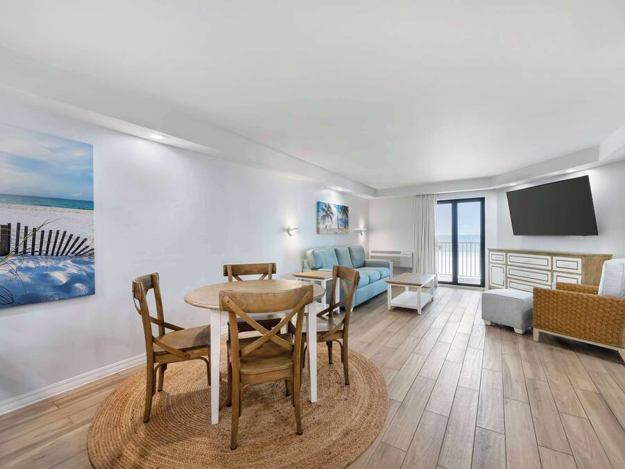 A modern living room with a round wooden table and chairs, a light blue sofa, a wicker armchair, and a flat-screen TV mounted on the wall.