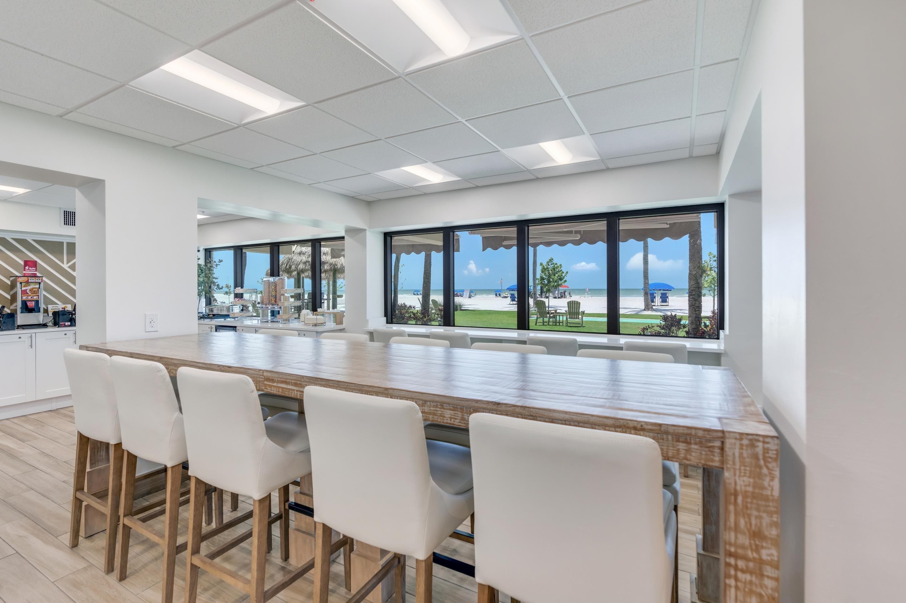 A modern kitchen with a large island, white bar stools, and large windows with a view of the beach.