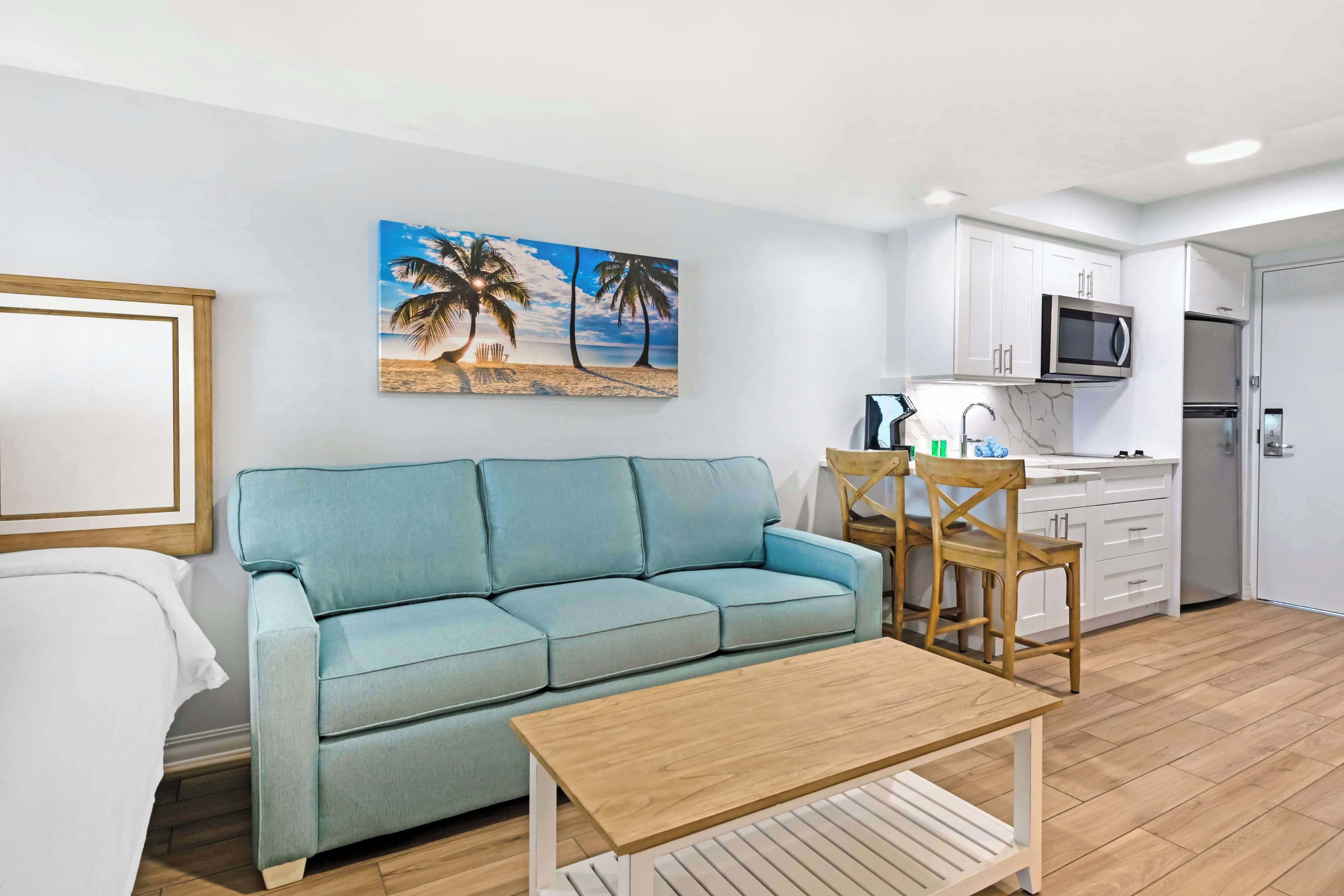 A living room with a sofa, a wooden coffee table, a dining area with a chair, and a kitchen with white cabinets and appliances.