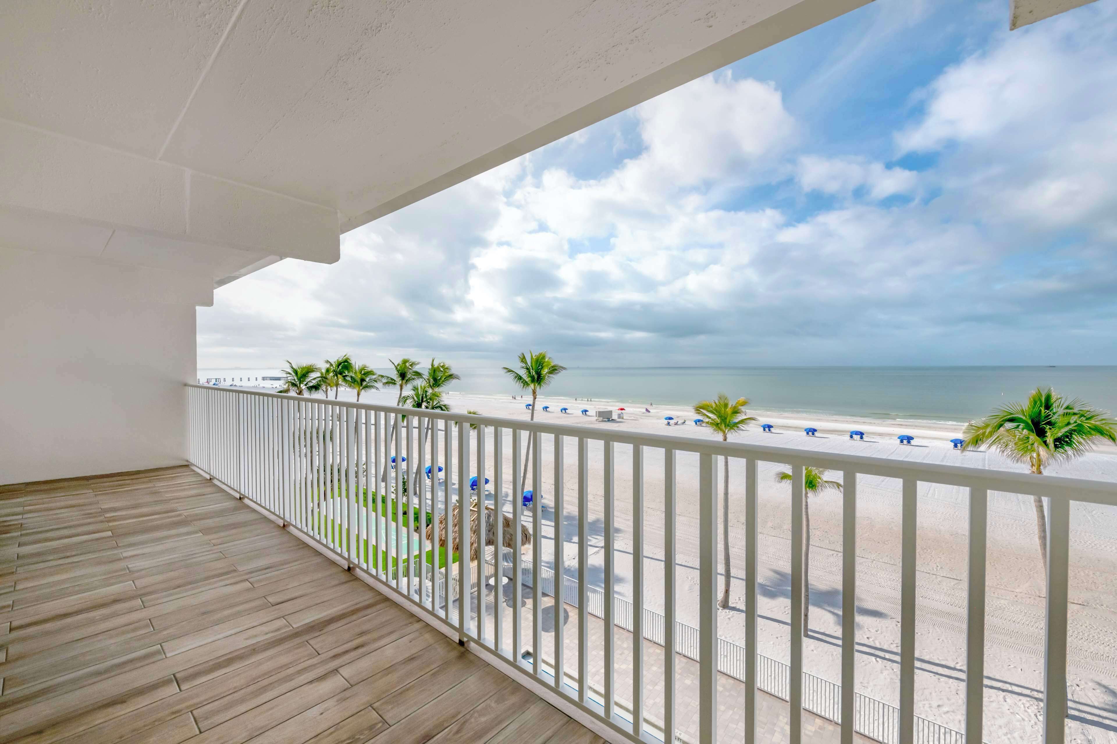 A balcony with a view of the beach, white railing, and wooden floor.