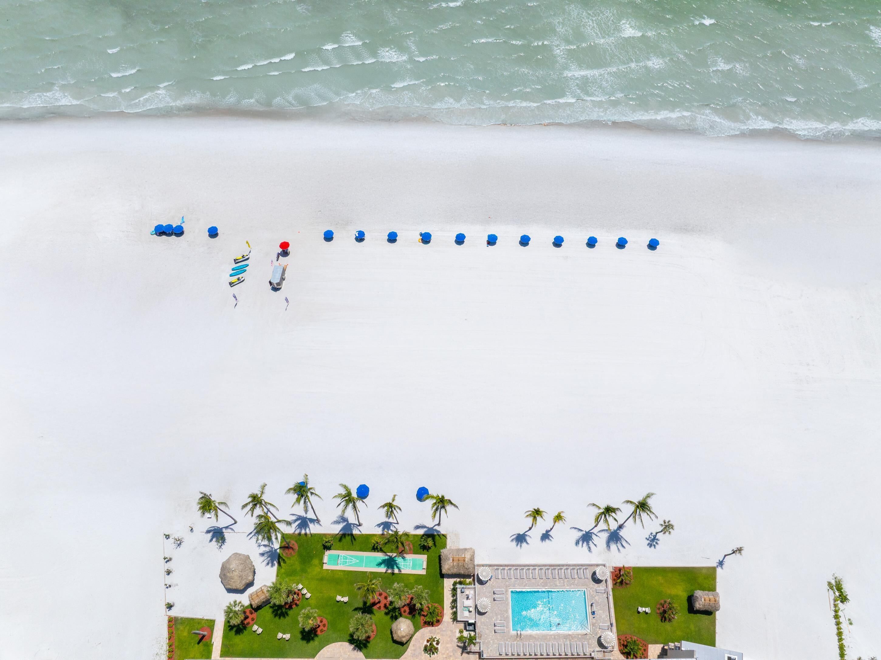 An aerial view of a beach resort with a swimming pool, lounge chairs, and palm trees.