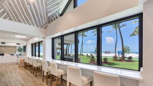A modern kitchen with white cabinets and a wooden floor, a bar area with white chairs, and large windows with a view of the beach and palm trees. - Best Western Plus Beach Resort (Fort Myers Beach)