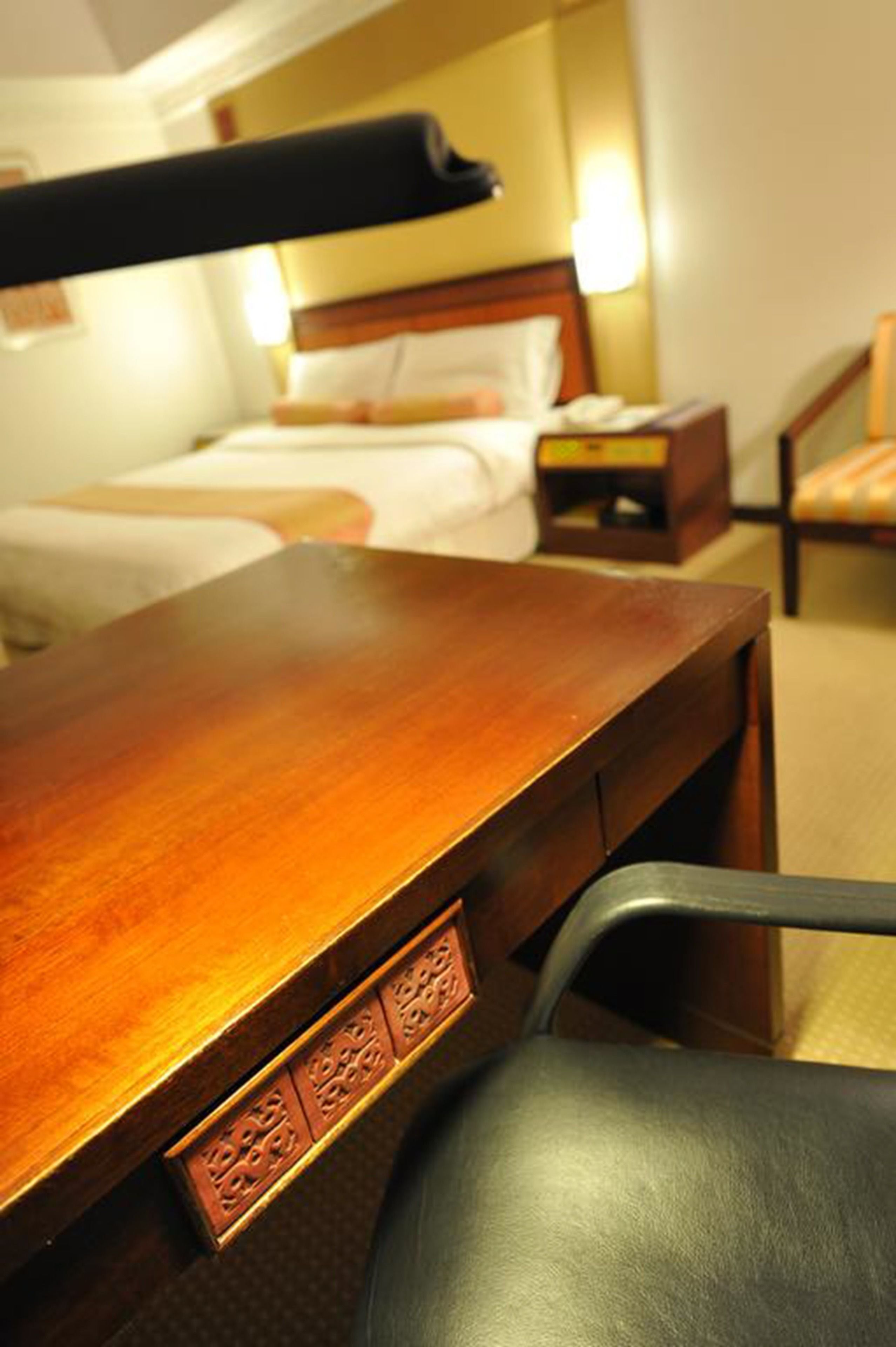 A hotel room with a wooden desk, a chair, a bed with white linens, a nightstand, and a lamp.