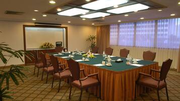Un salón de conferencias con una larga mesa, sillas, una pantalla de proyector y una planta.