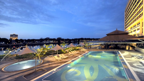 A swimming pool with lounge chairs and umbrellas, surrounded by a building with a view of a city skyline. - Grand Margherita Hotel (Kuching)