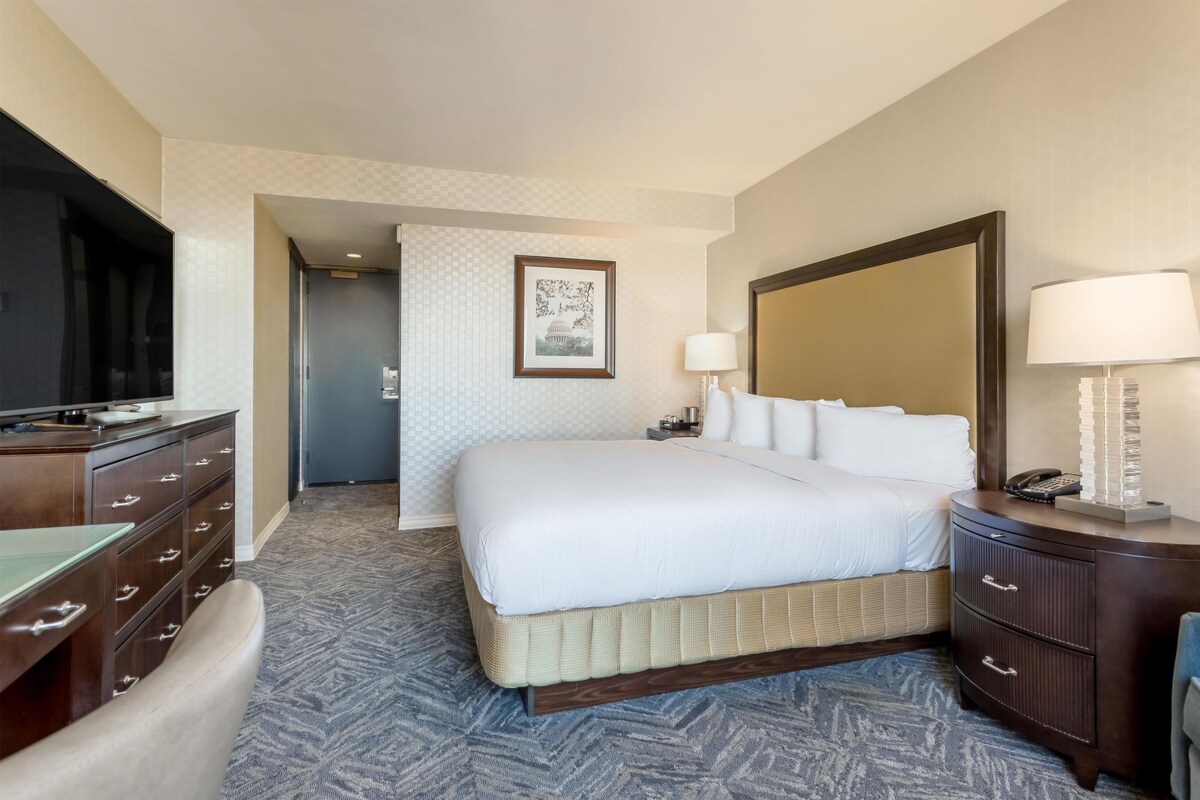 A hotel room with a bed, a television, a desk with drawers, a lamp, and a framed picture on the wall.