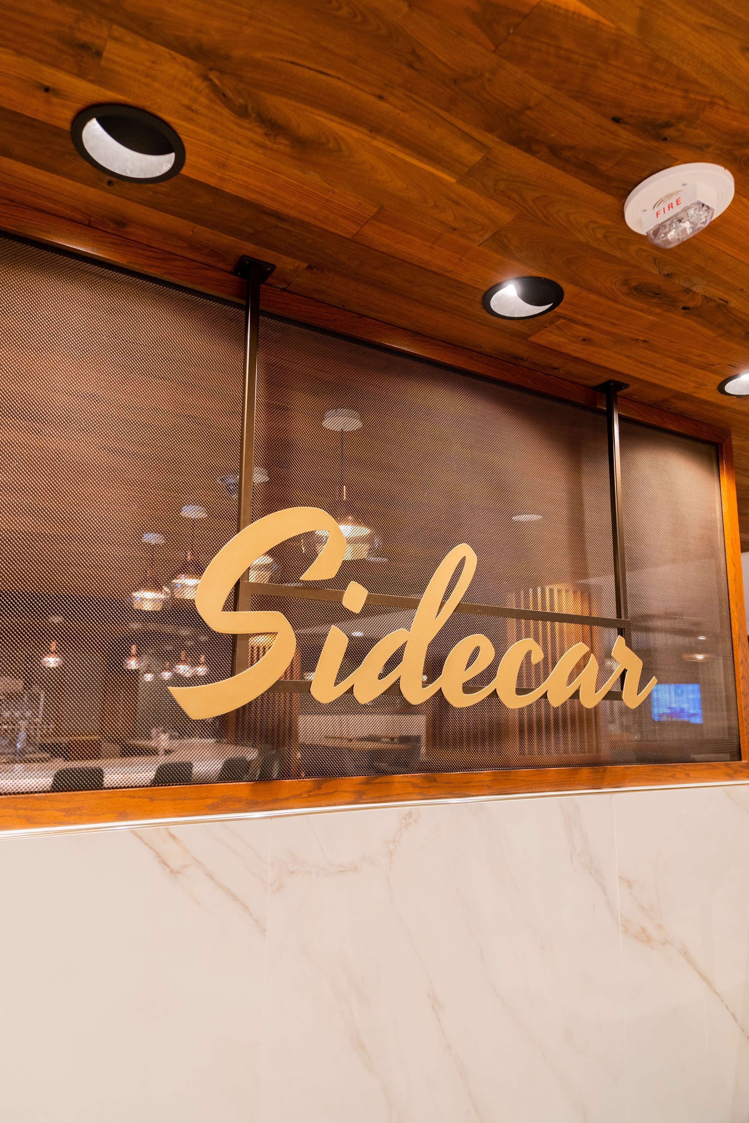Une enseigne portant le mot « Sidecar » est fixée à un mur doté d’un plafond en bois et d’un éclairage encastré.