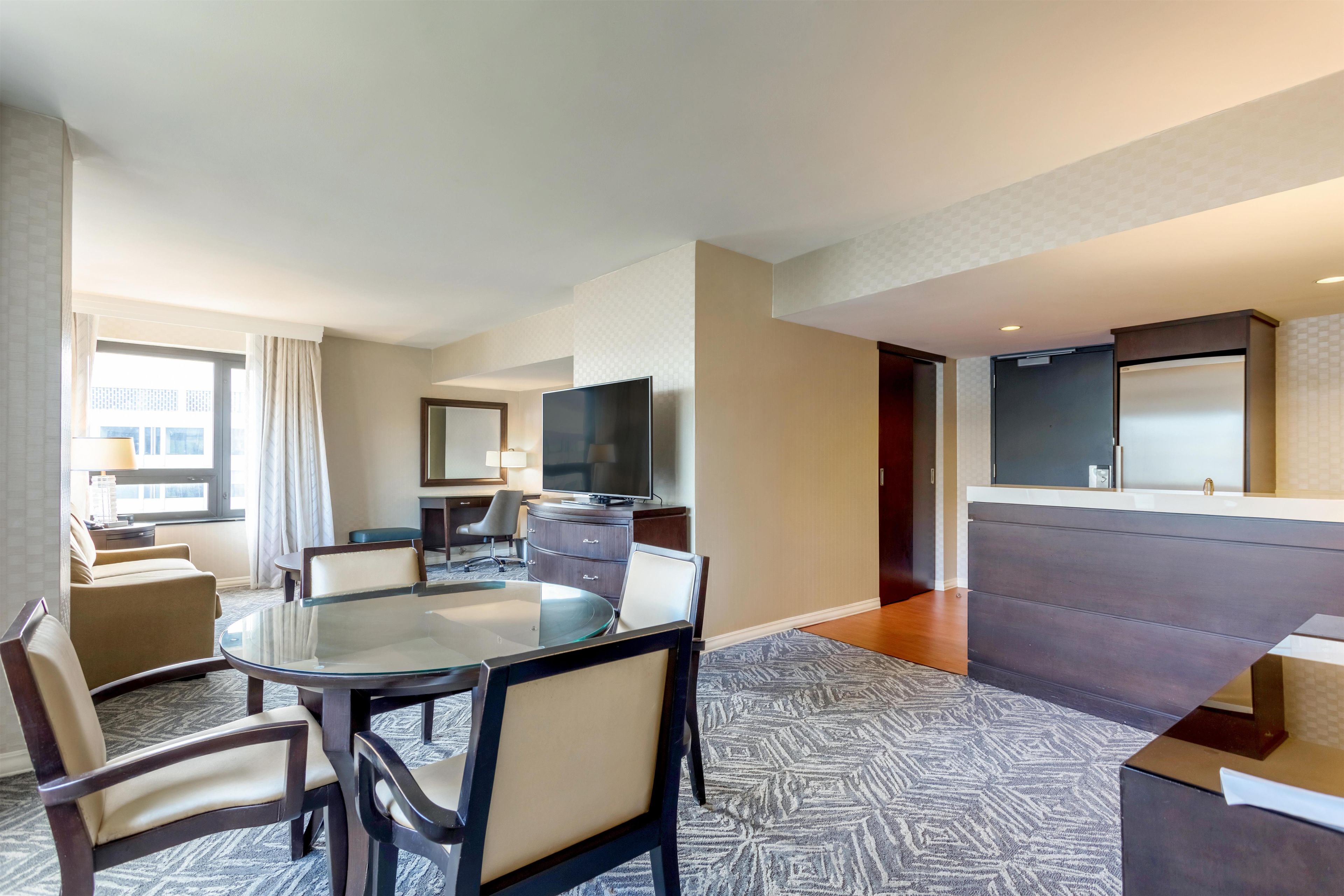 A hotel room with a dining area, a TV, a sofa, and a large window.
