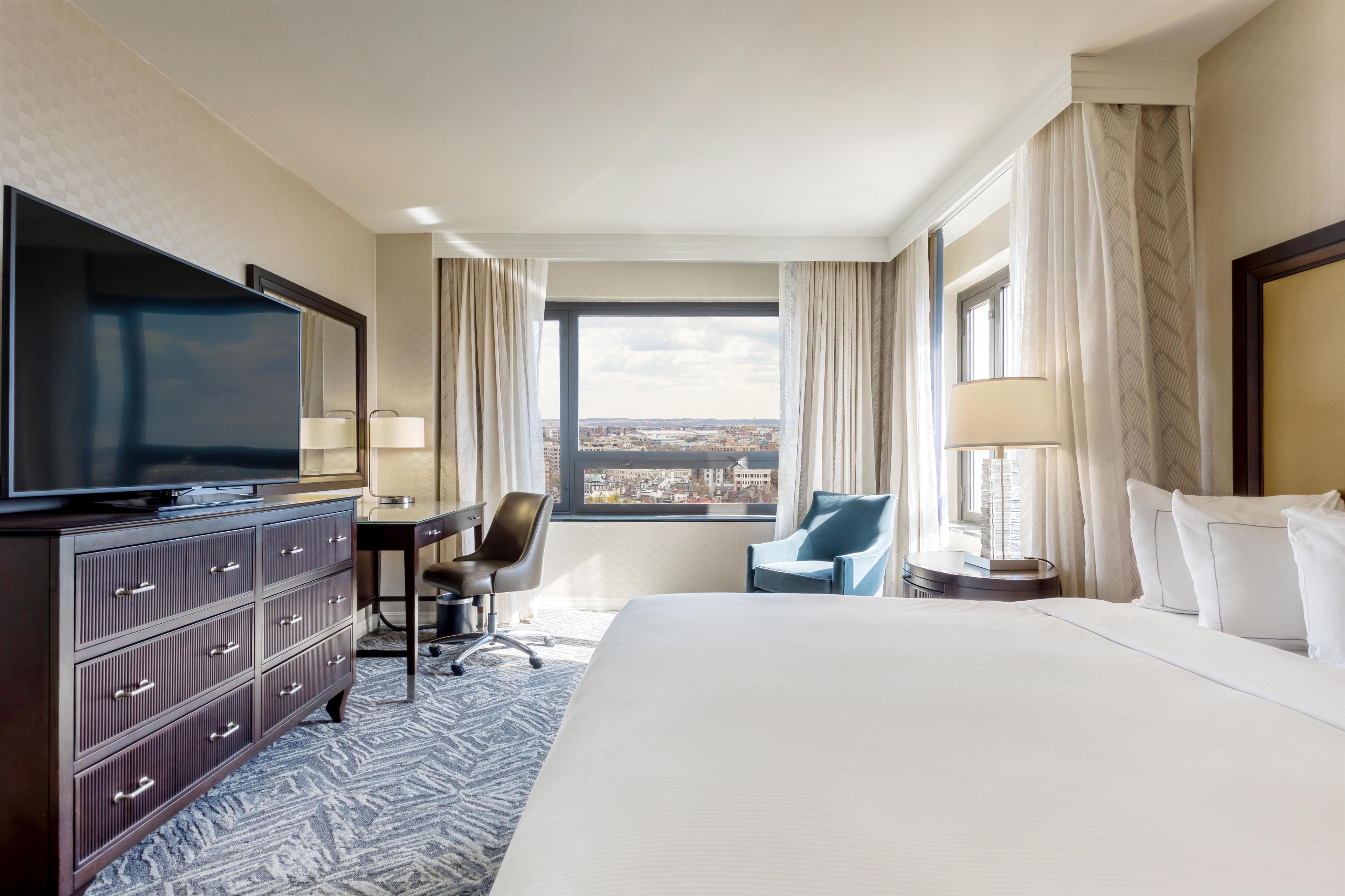 Habitación de hotel con una cama grande, un escritorio, una silla, televisión y vista de la ciudad.