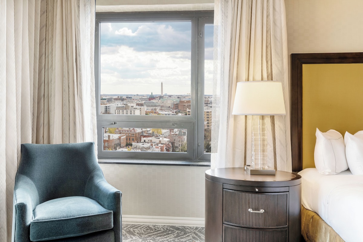 A hotel room with a large window, a bed, a chair, a nightstand, and a lamp.