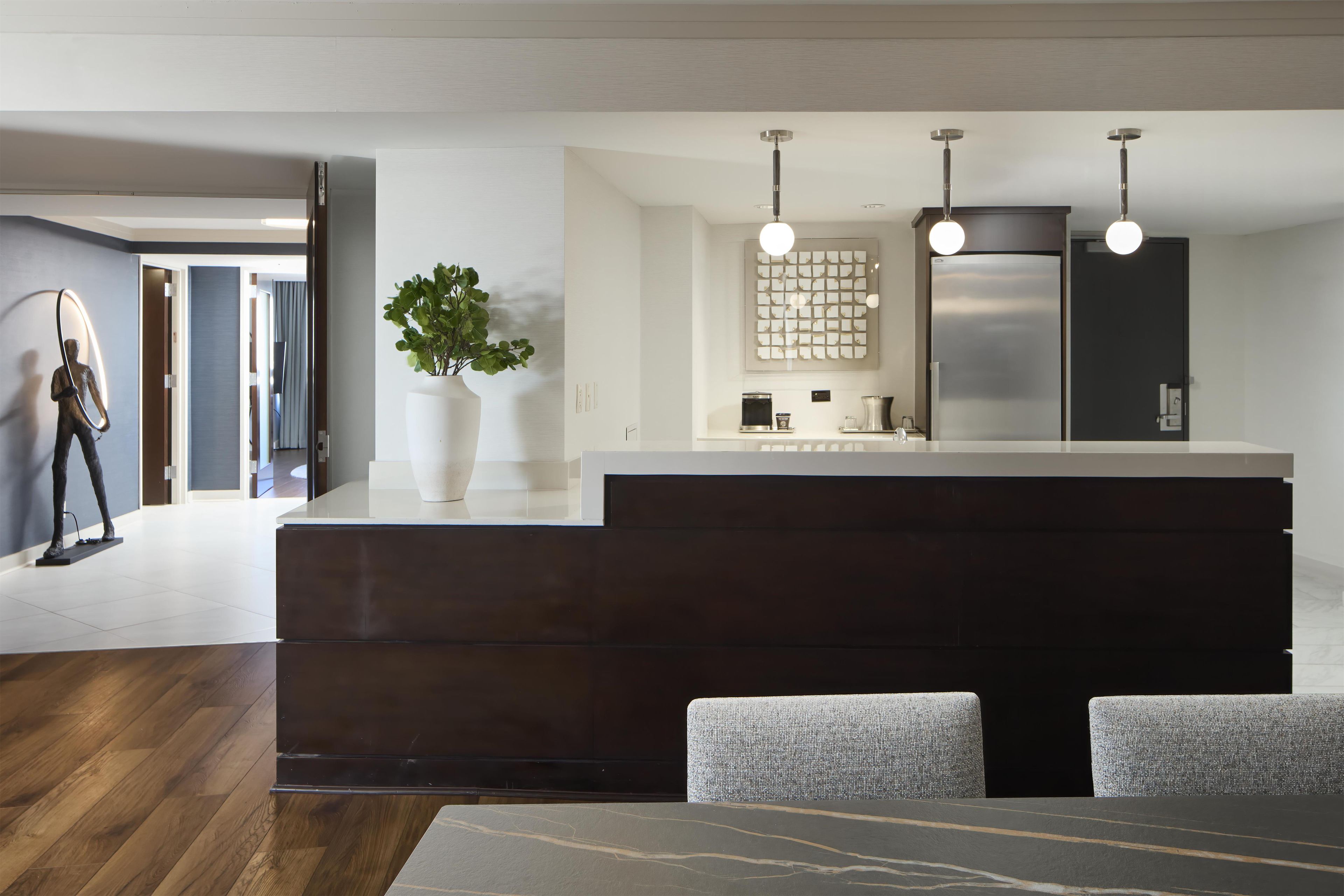 A modern kitchen with a dark wood island, white countertops, and stainless steel appliances. There is a plant on the island and a seating area with a gray upholstered bench.