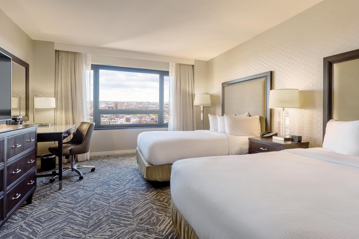 A hotel room with two beds, a desk, a chair, and a large window offering a city view.