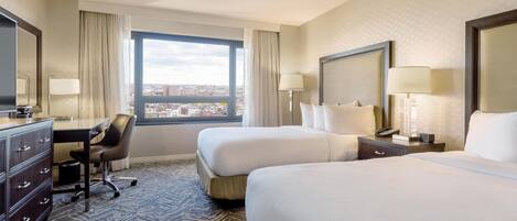 Habitación de hotel con dos camas, un escritorio, una silla y un amplio ventanal con vistas a la ciudad.