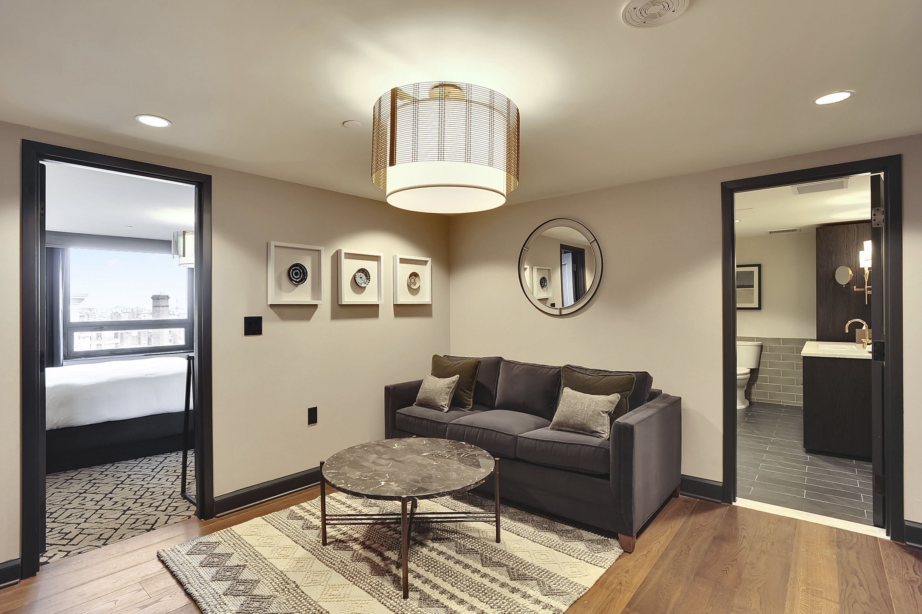 A modern living room with a dark grey sofa, a round coffee table, and a large mirror on the wall. There are framed pictures on the wall and a ceiling light fixture.