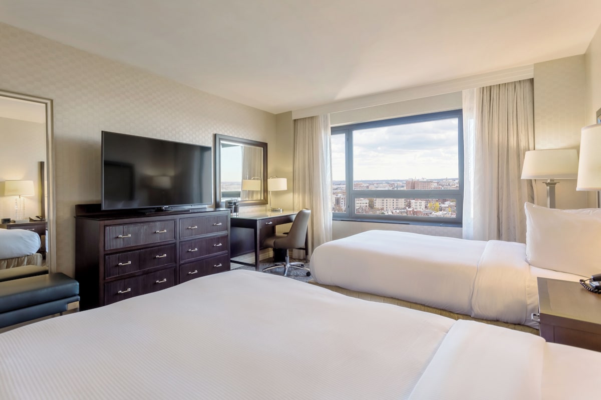 A hotel room with two beds, a TV, a desk, and a view of the city.