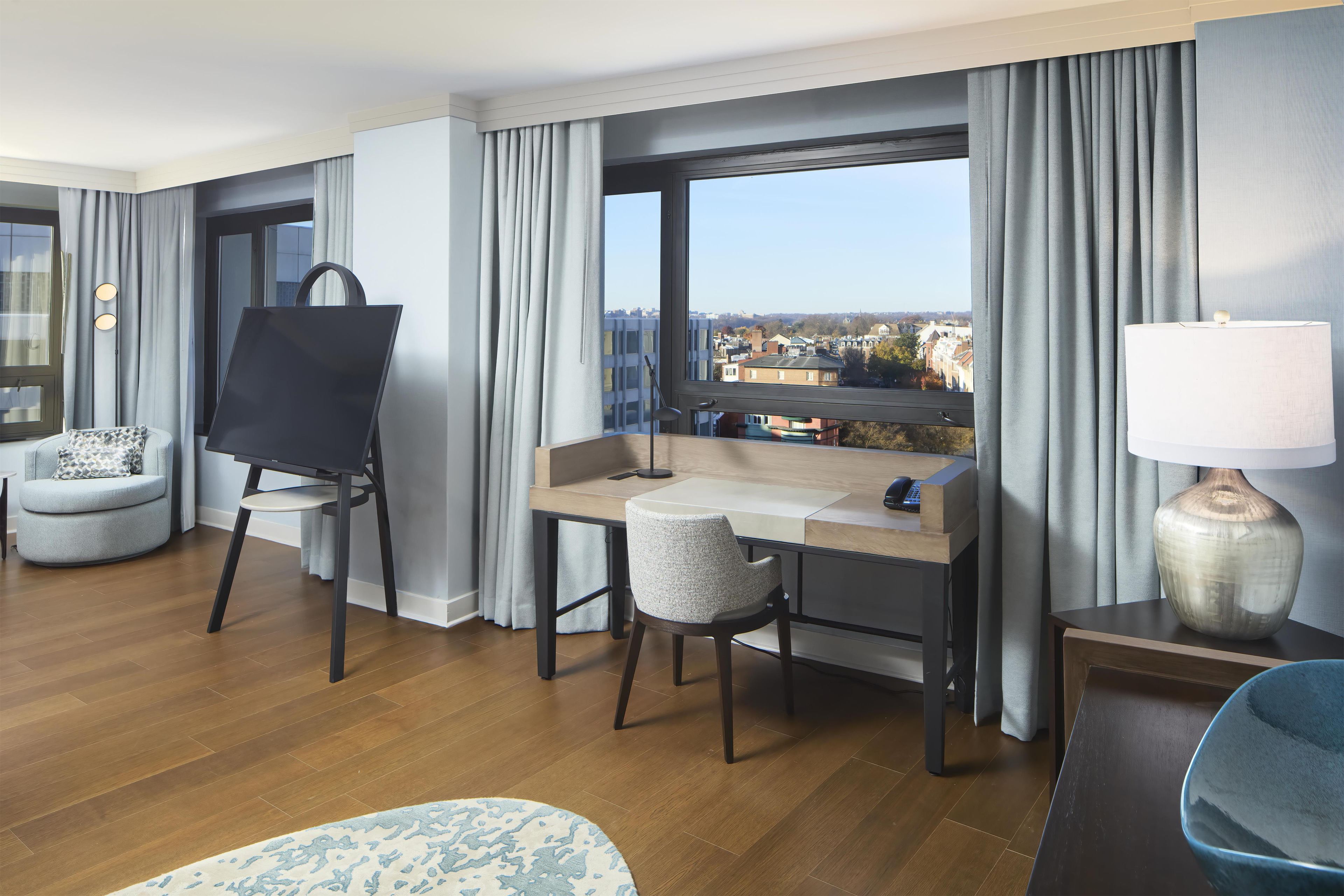 A hotel room with a desk, a chair, an easel, a lamp, and a view of the city through the window.