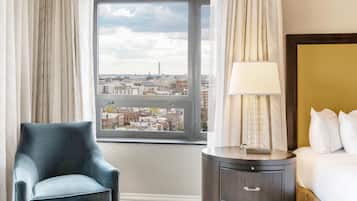 A hotel room with a view of the city, a bed, a nightstand with a lamp, and a blue armchair.