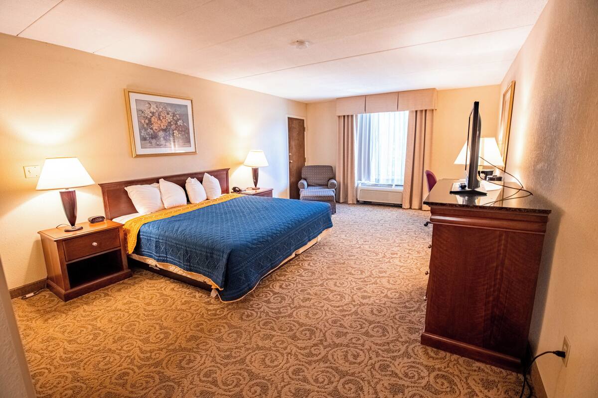 A hotel room with a large bed, two bedside tables with lamps, a desk with a computer, a chair, and a window with curtains.