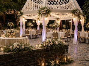An outdoor wedding reception area with round tables, white drapery, floral arrangements, and string lights.