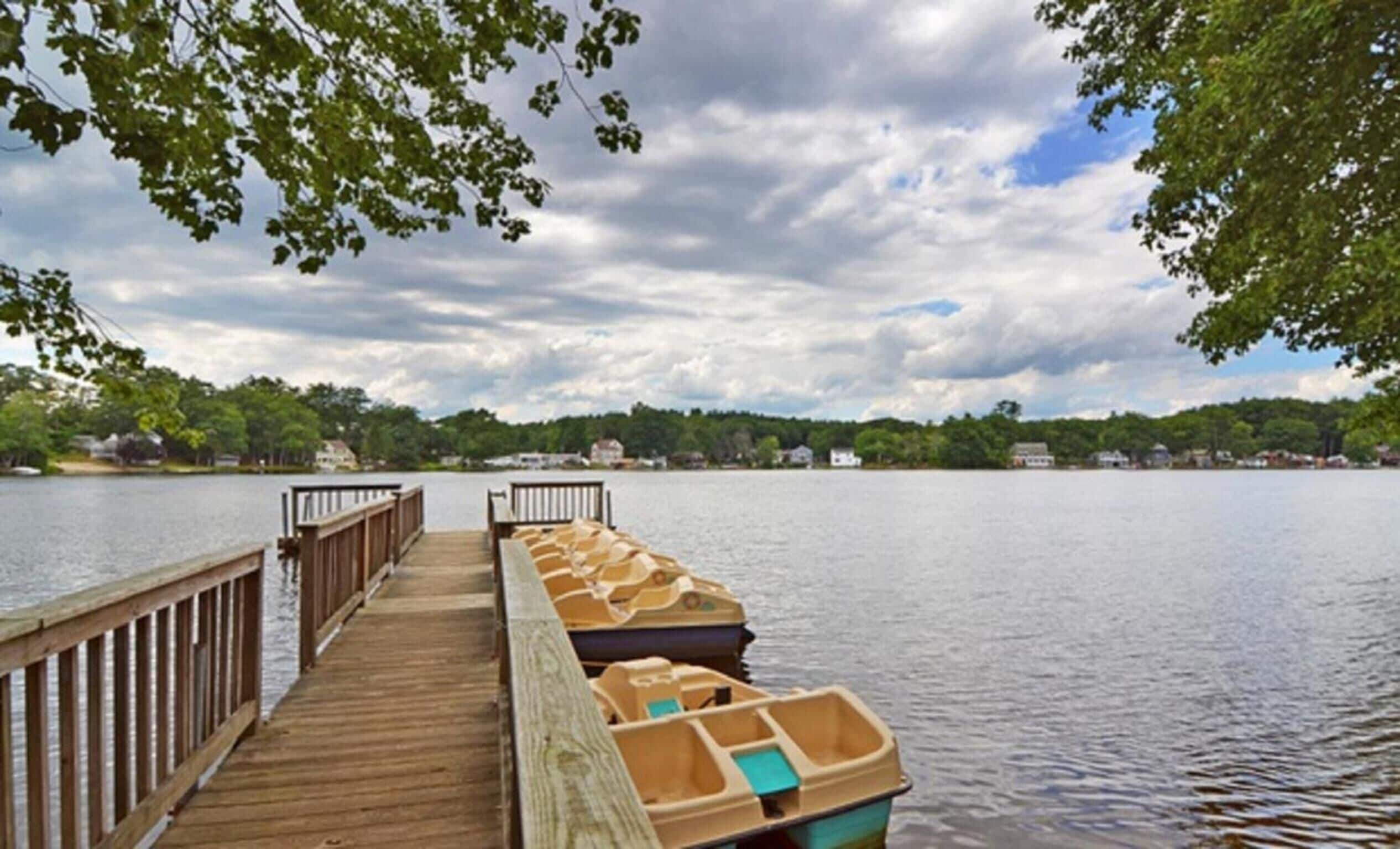 A wooden dock leading to a row of small boats on calm water, surrounded by trees and houses in the distance.