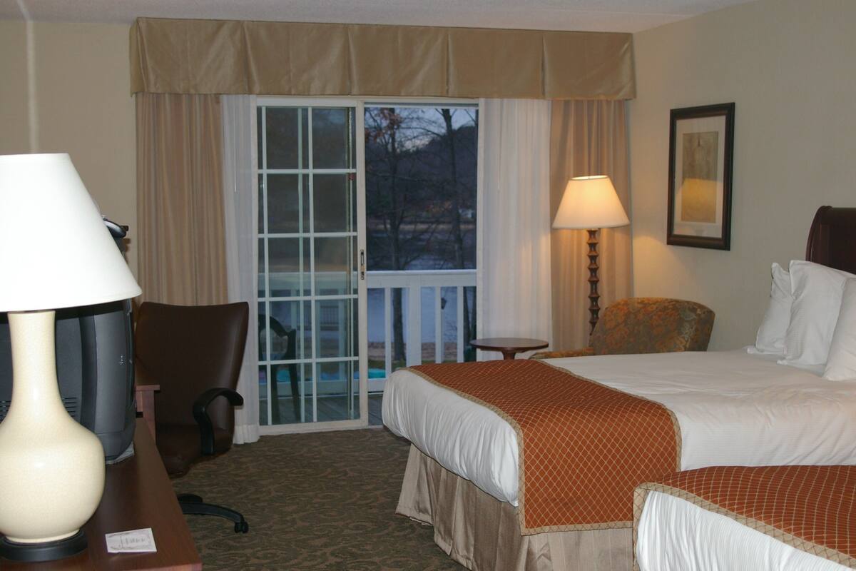A hotel room with a bed, a chair, a desk with a lamp, a small table, and a window with curtains.