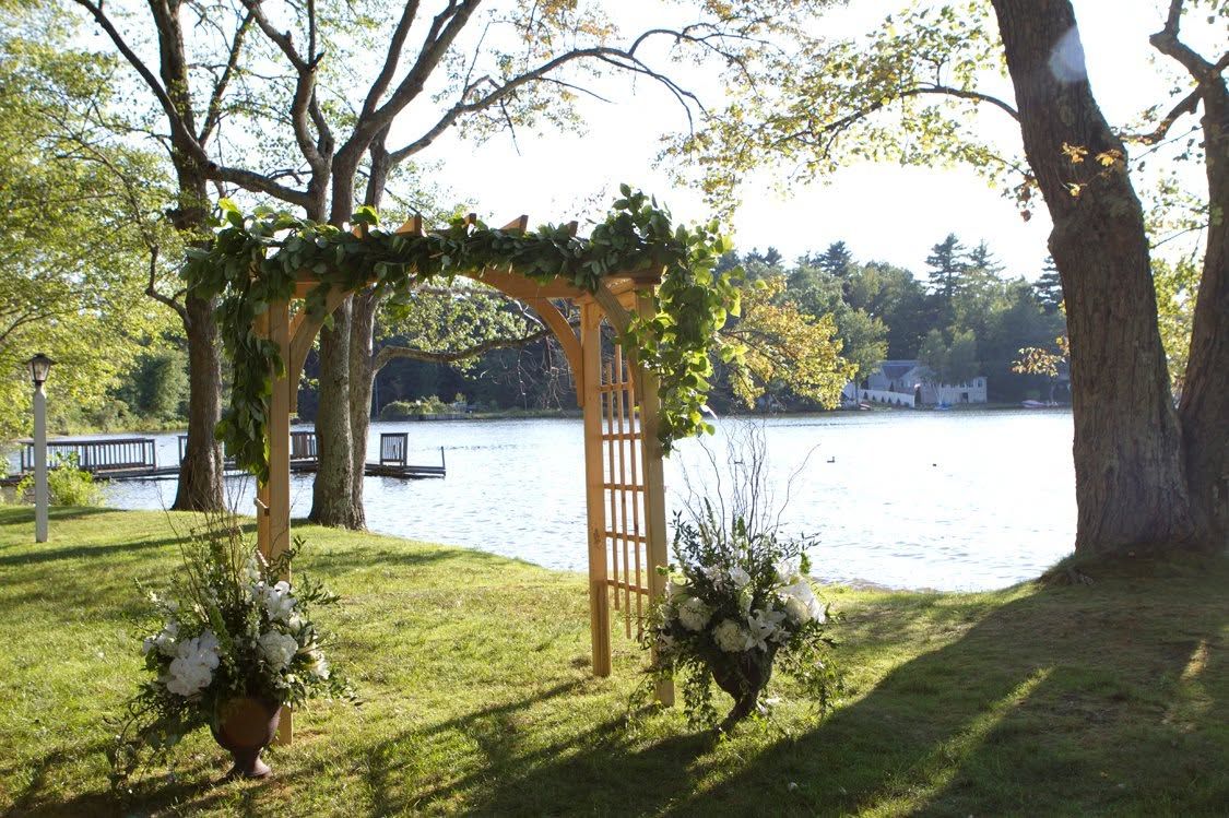 A gazebo with a wooden frame and a vine-covered arch, surrounded by grass and trees, with a body of water and a dock in the background.