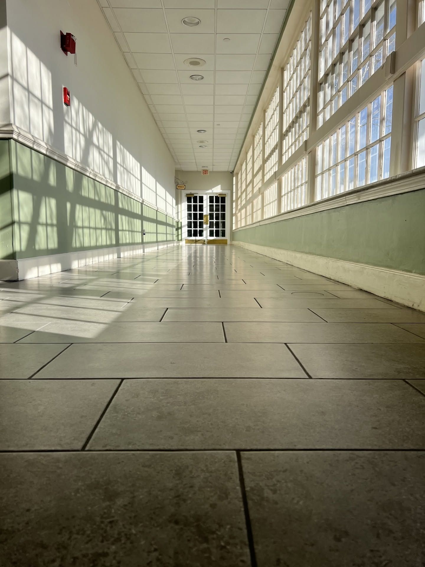 A long, empty corridor with tiled flooring and large windows on one side.