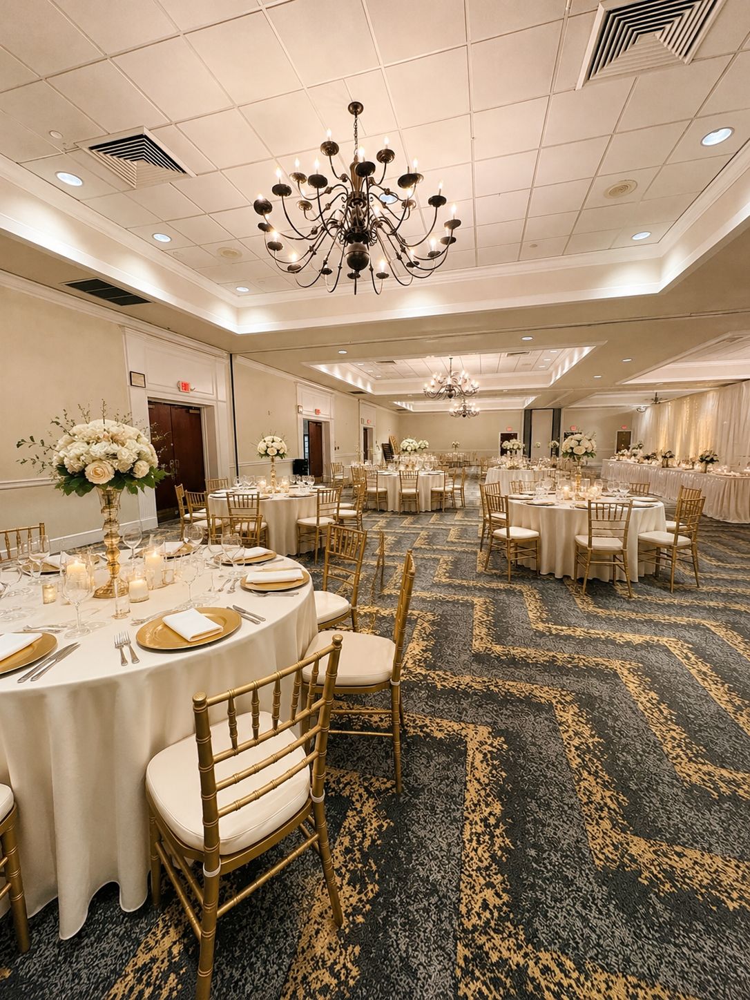 A banquet hall with round tables, gold chairs, and chandeliers.