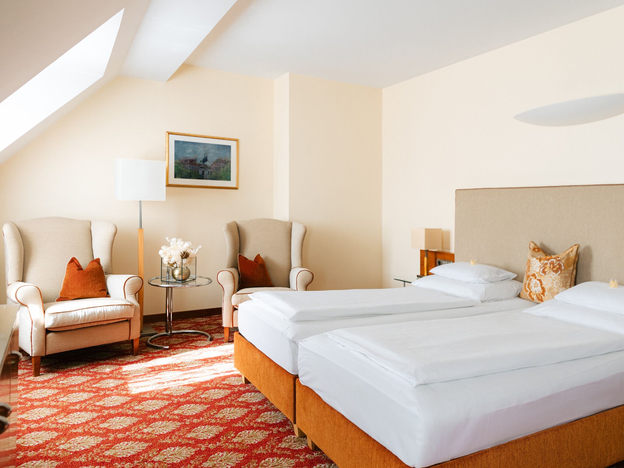 A hotel room with two beds, a sofa, a small table with a vase of flowers, a lamp, and a painting on the wall.