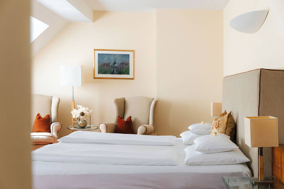 A hotel room with a large bed, two armchairs, a small table with a vase of flowers, a painting on the wall, and a lamp on the nightstand.
