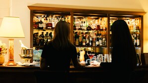 Dos personas sentadas en un bar con una barra de licores bien surtida al fondo.