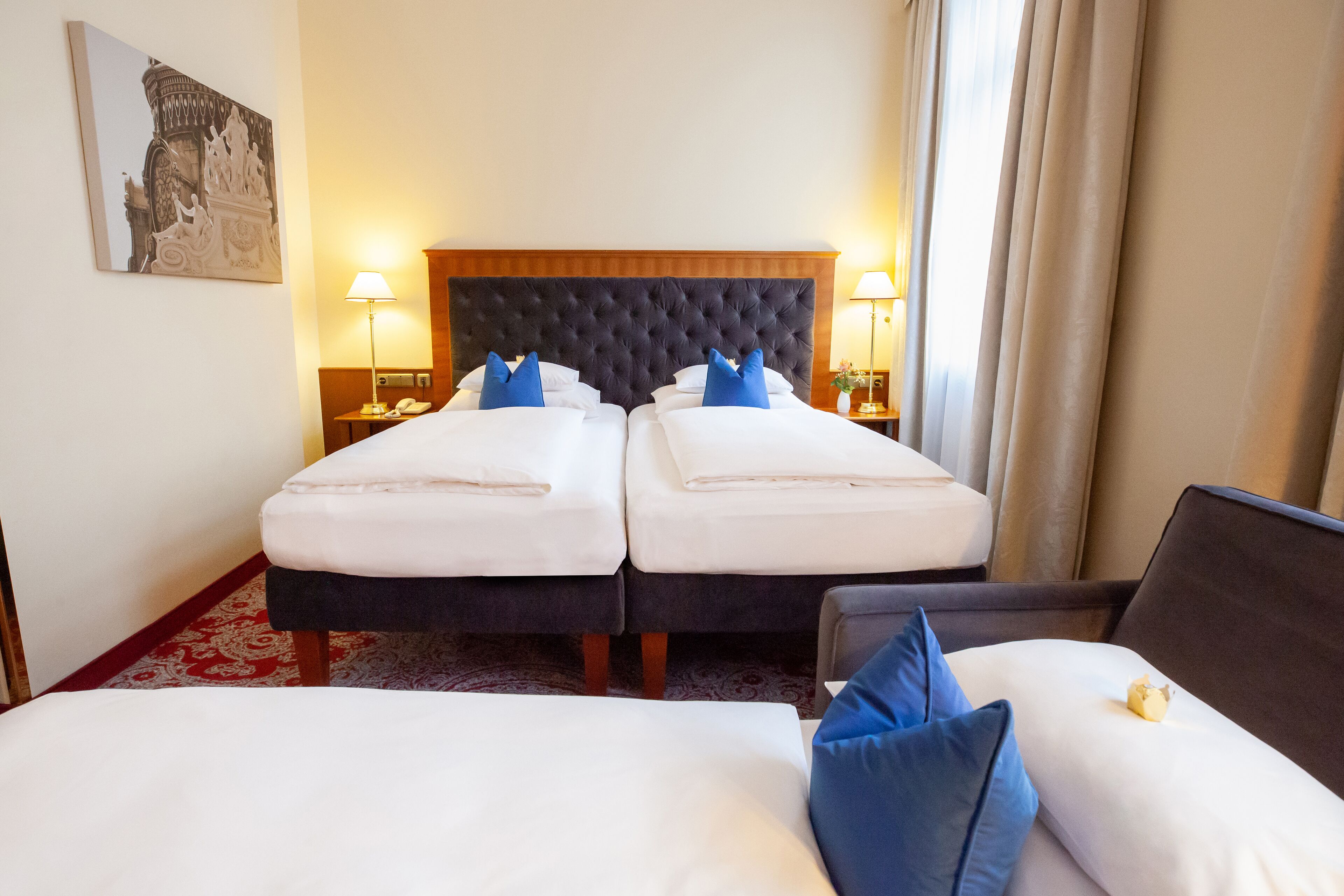 Quarto de hotel com duas camas, cabeceira grande, luminárias de cabeceira e um detalhe decorativo na parede.