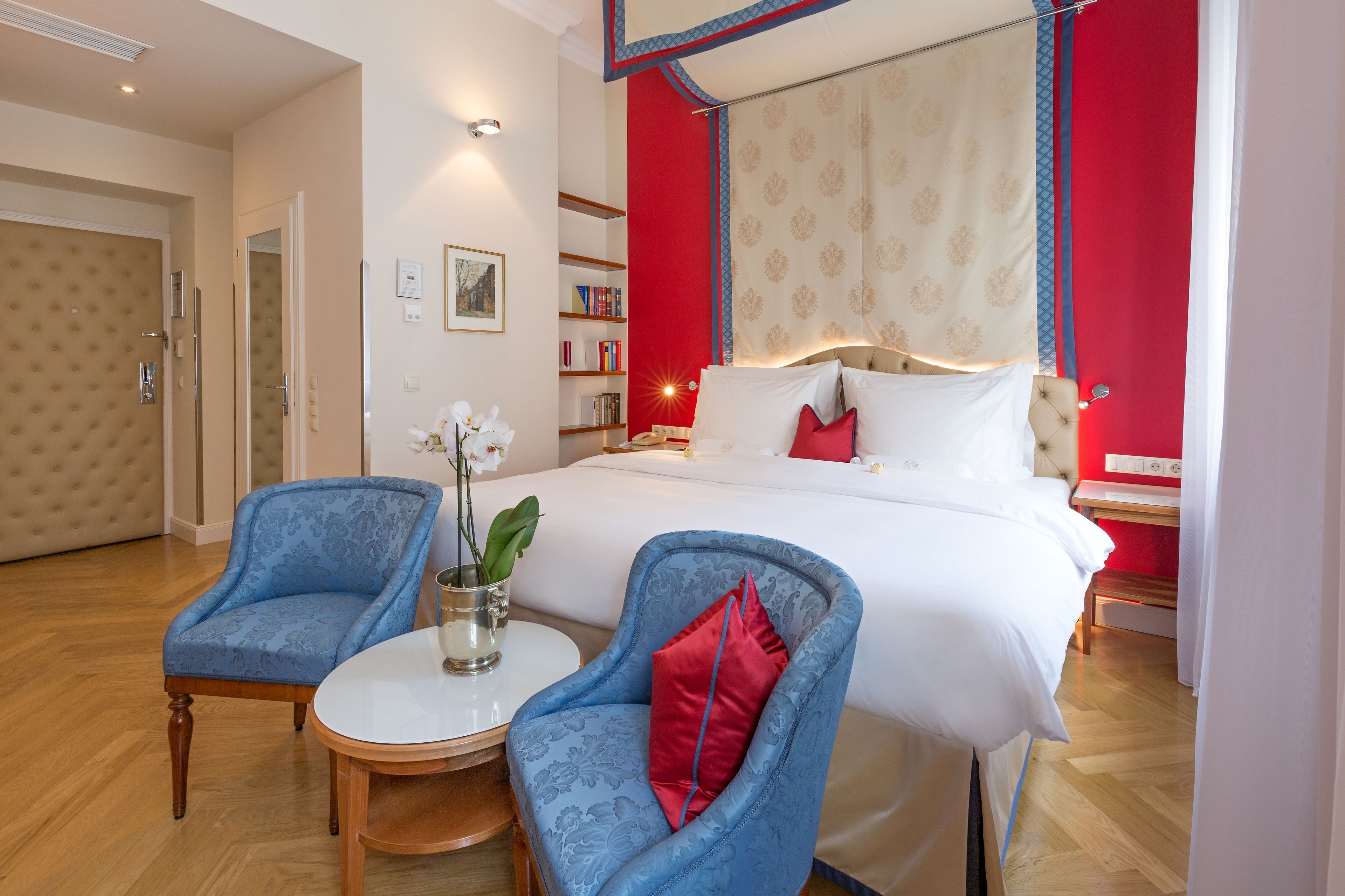 A hotel room with a large bed, two blue armchairs, a small round table, and a bookshelf.