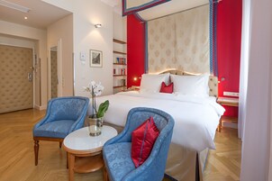 Quarto de hotel com uma cama grande, duas poltronas azuis, uma mesinha redonda e uma estante de livros.