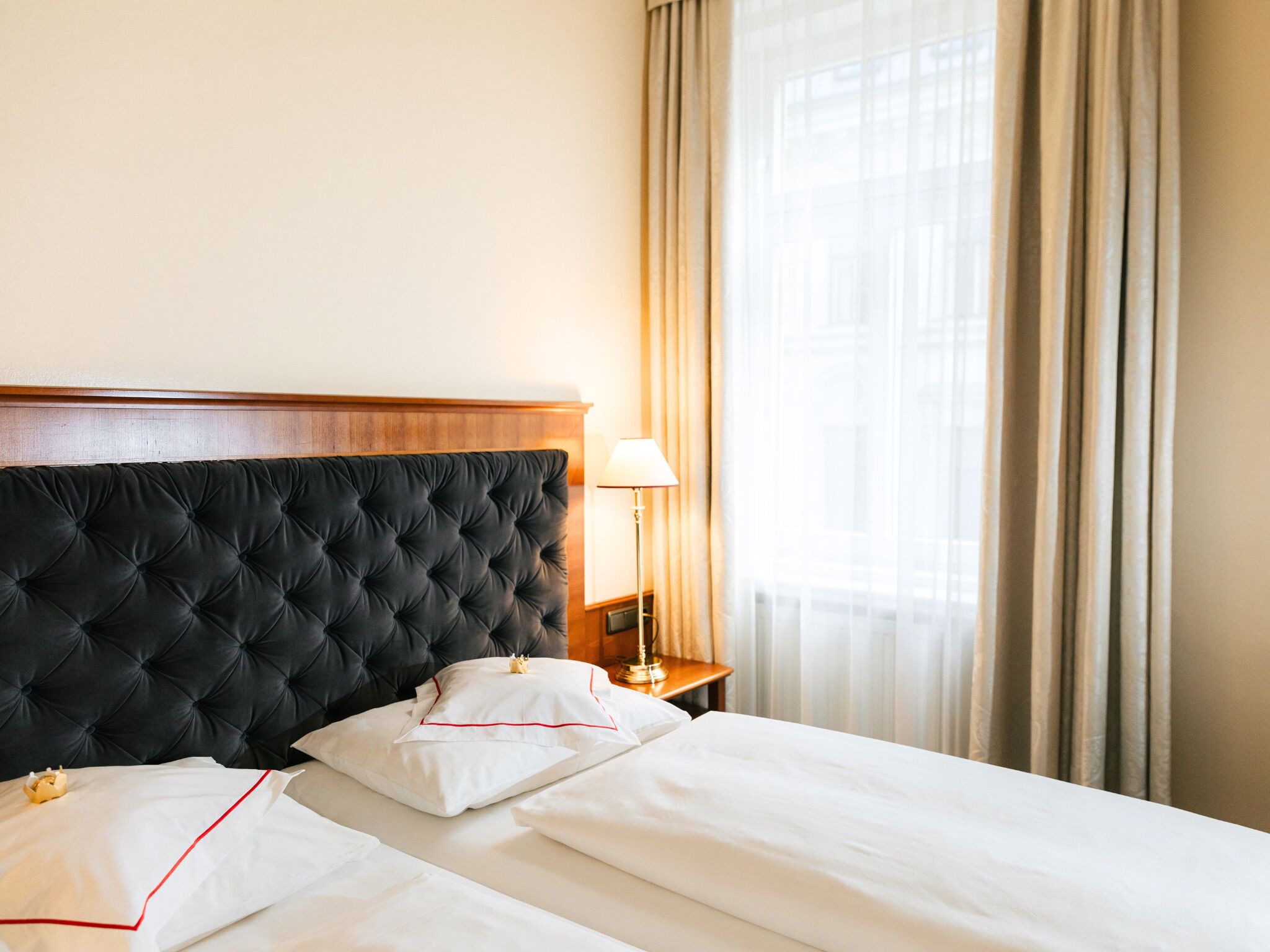Quarto de hotel com cabeceira estofada, luminária ao lado da cama e uma janela grande com cortinas.