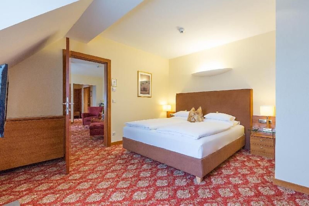 A hotel room with a large bed, a wooden headboard, a red patterned carpet, a wooden nightstand, and a door leading to another room.