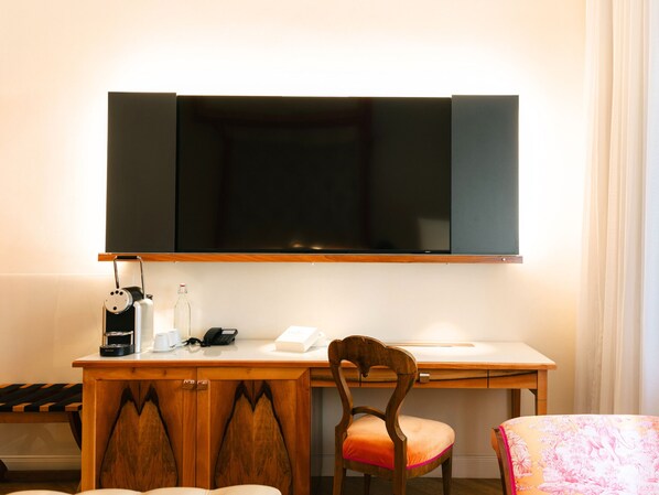 A hotel room with a desk, a chair, a television mounted on the wall, a coffee maker, a bottle, and a telephone.