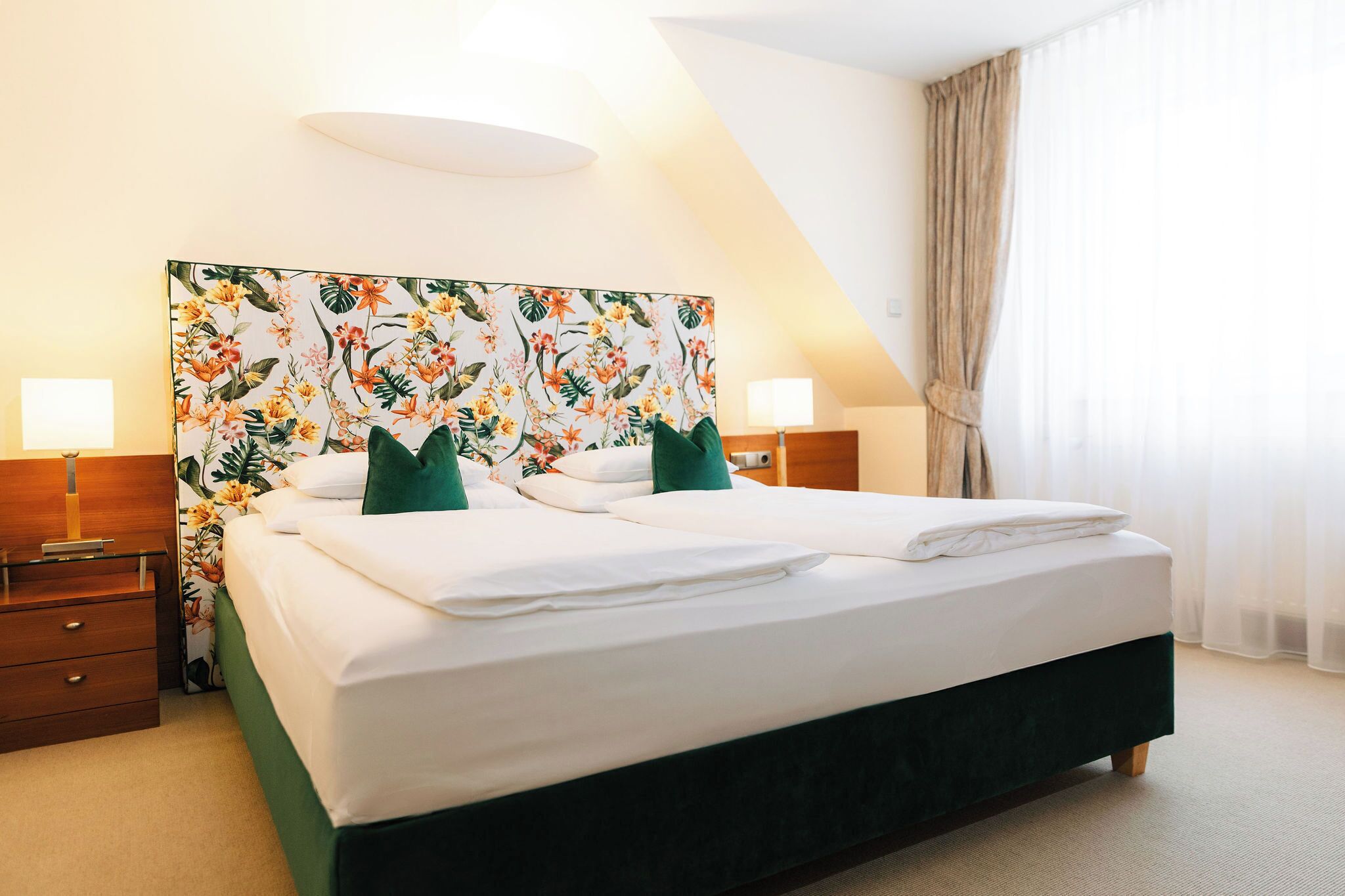 Una camera d'albergo con un letto grande, due comodini, una testiera fiorata e una finestra con tende.