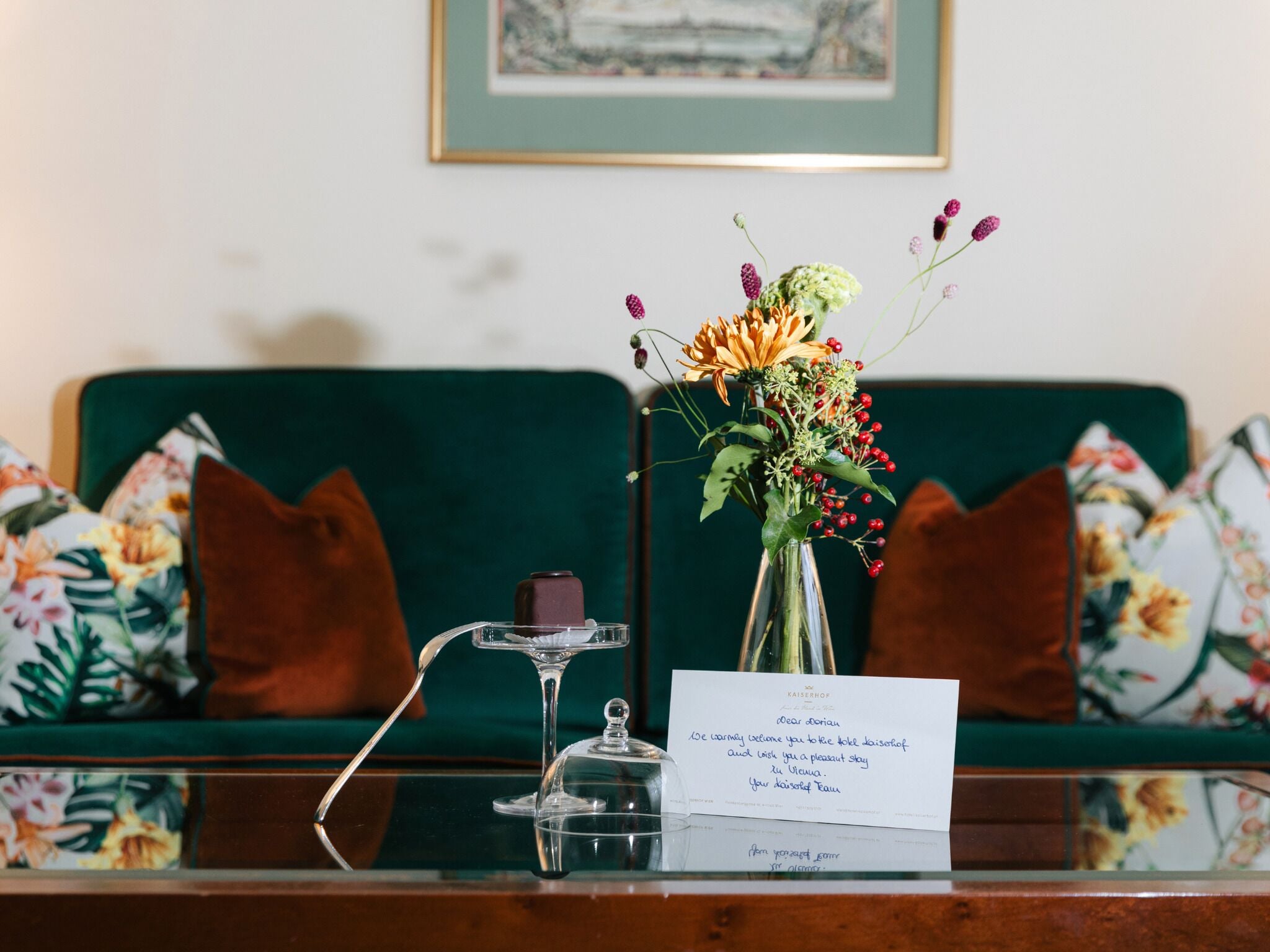 A green sofa with floral cushions, a clear vase with flowers, a glass candle holder, and a card on a glass table.