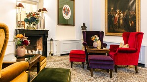 Una habitación con chimenea, sillones rojo y morado, una mesa con un florero y un cuadro grande en la pared.