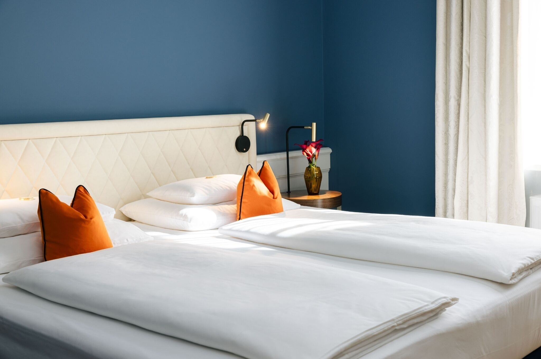A neatly made bed with white linens and orange pillows, a bedside table with a vase and a lamp, and a blue wall.