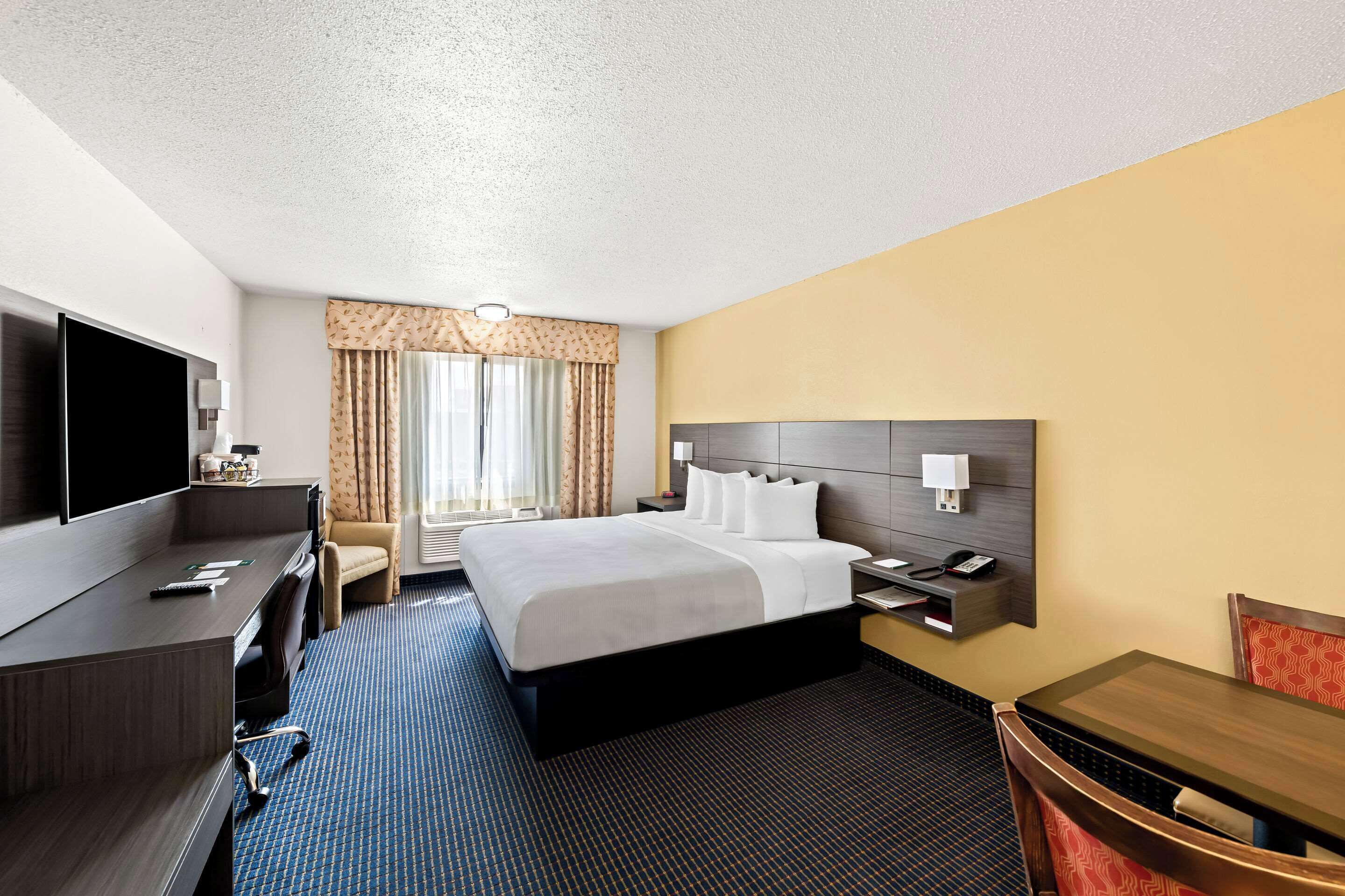 A hotel room with a large bed, a desk with a flat-screen TV, a chair, a table, and a window with curtains.