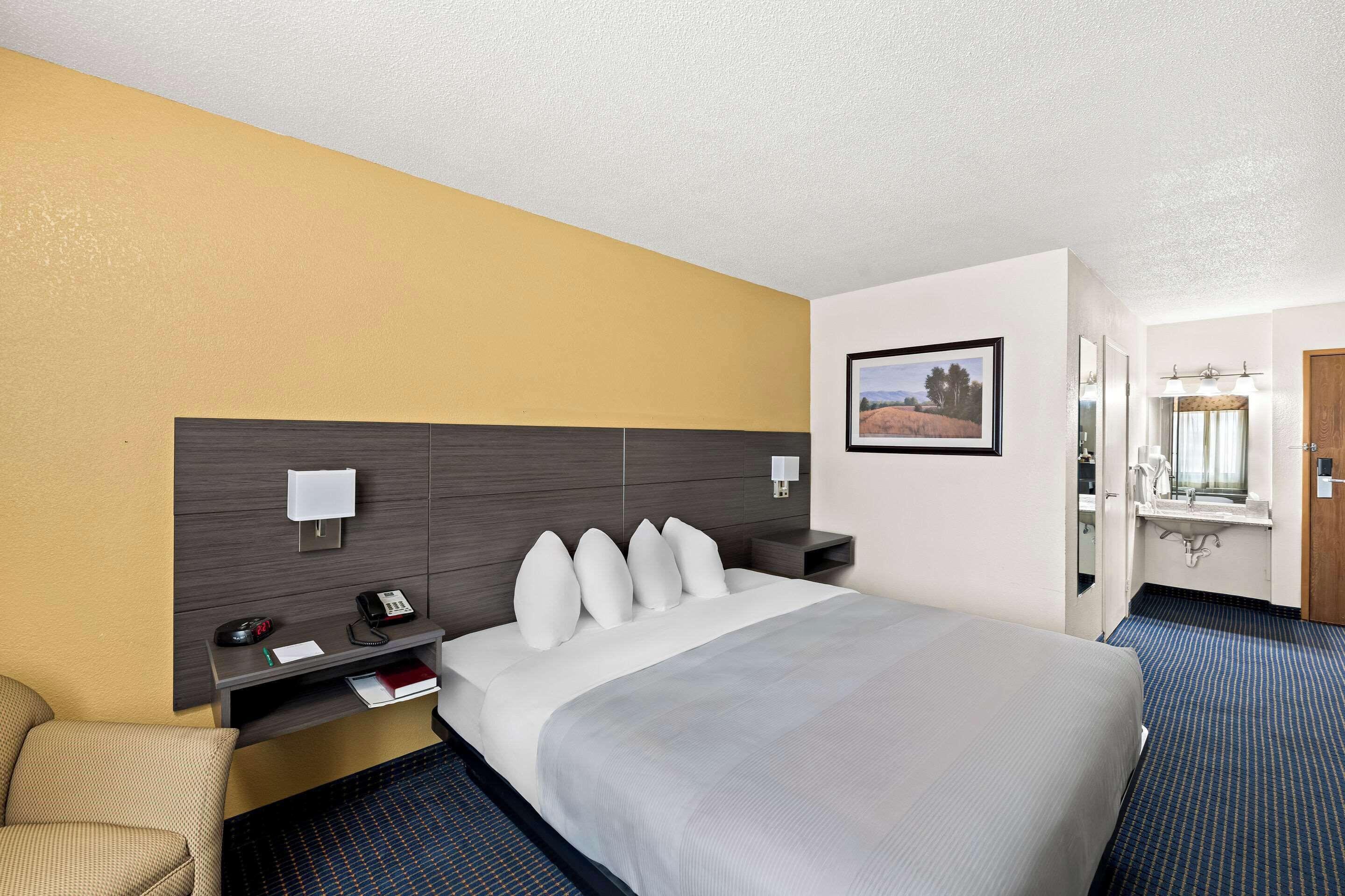 Una habitación de hotel con una cama grande, una mesita de noche con lámpara, un escritorio con teléfono, y un baño visible a través de una puerta abierta.