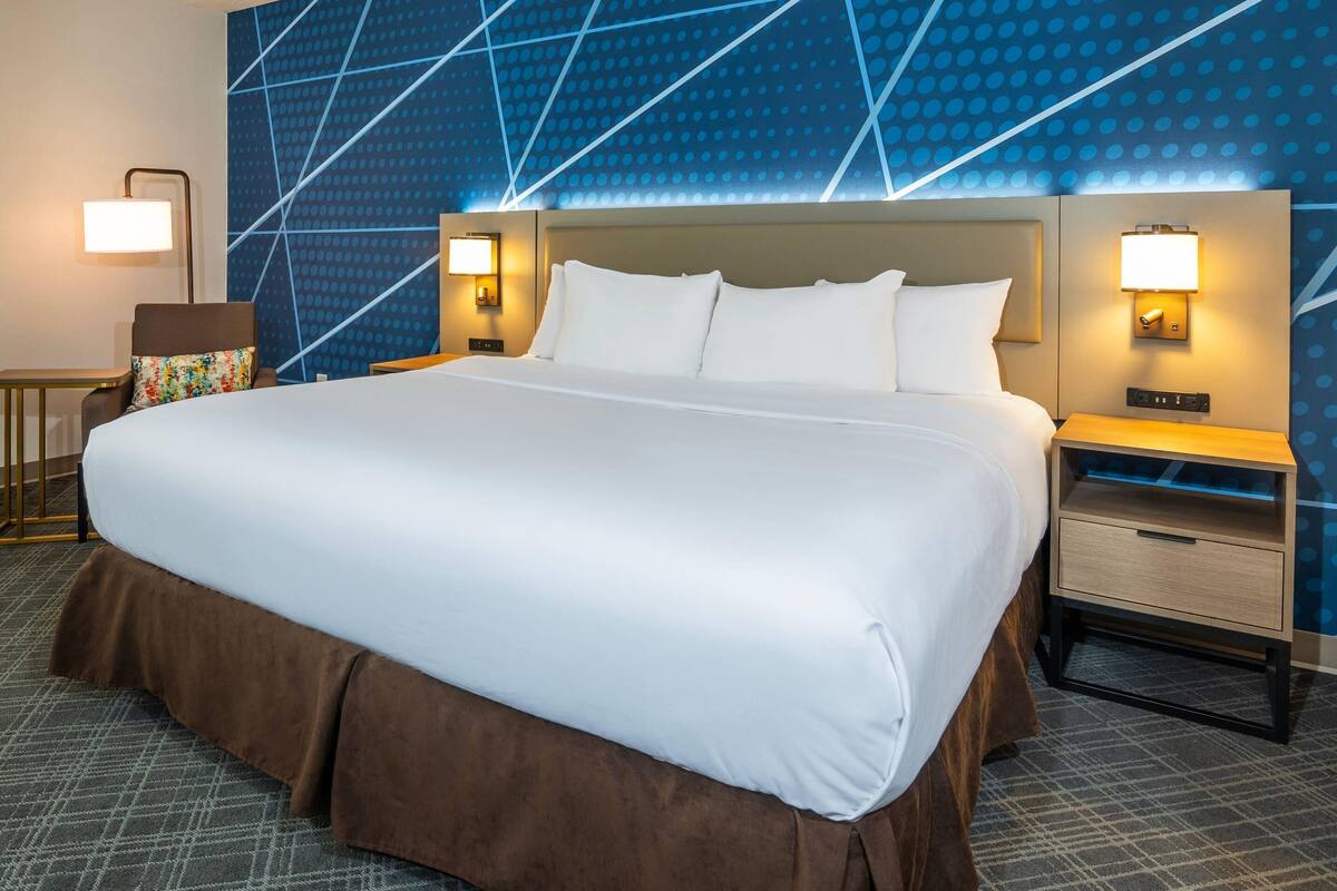 A hotel room with a large bed, two bedside tables with lamps, a chair, and a patterned wall.