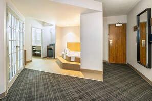 A hotel hallway with a carpeted floor, a wooden door, a mirror, and a bathroom with a bathtub and sink.
