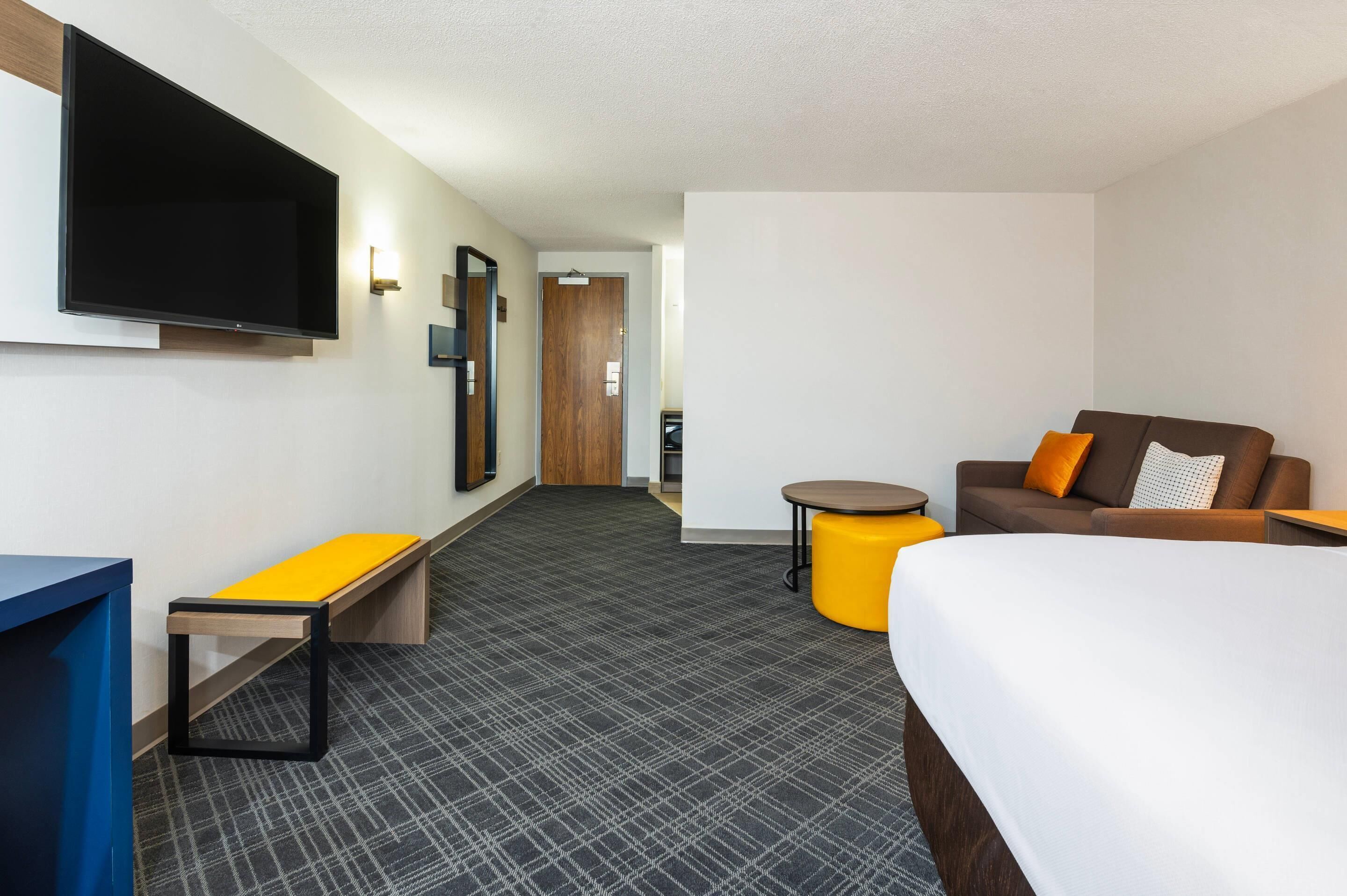 A hotel room with a bed, a sofa, a small table, a bench, and a TV mounted on the wall.